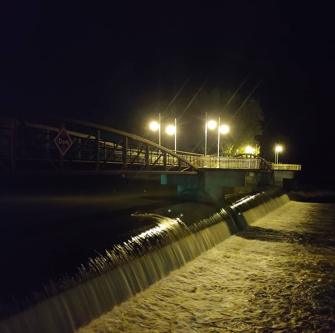 a bridge at night, crossing over a squat, rushing dam and lit only by its few lampposts 